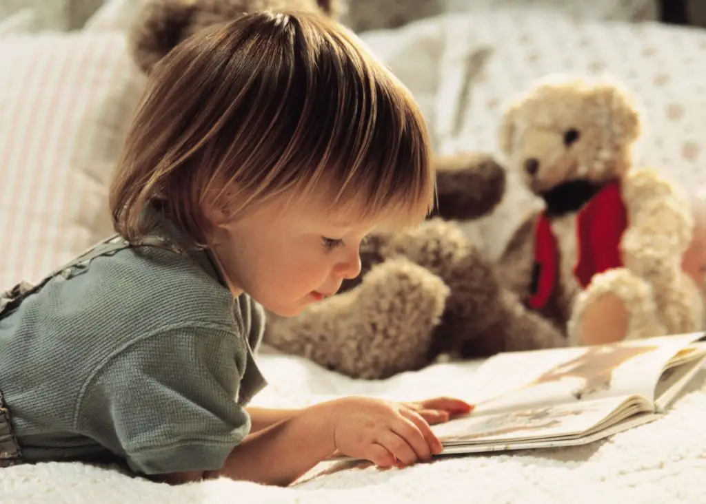 3 Year Old Schedule No Nap: toddler laying on belly on bed reading a book quietly with cute teddy bear in the background